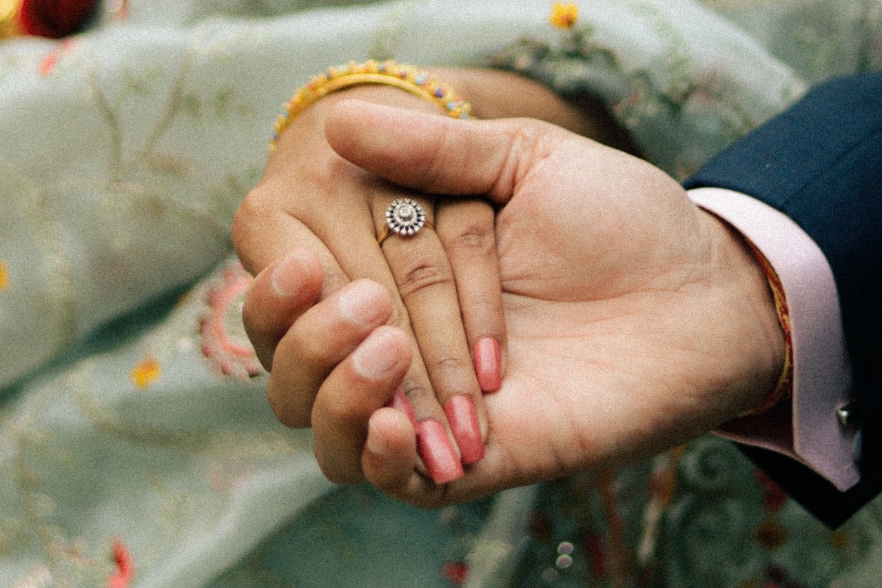 A close-up of a couple’s hands, the woman wearing an elegant halo engagement ring.