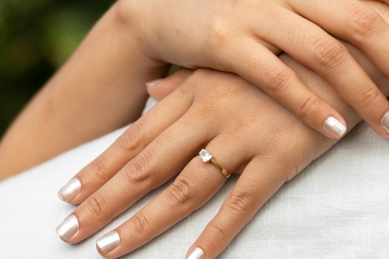 A close-up of a bride-to-be’s hands, wearing a solitaire engagement ring as she embraces her fiance.