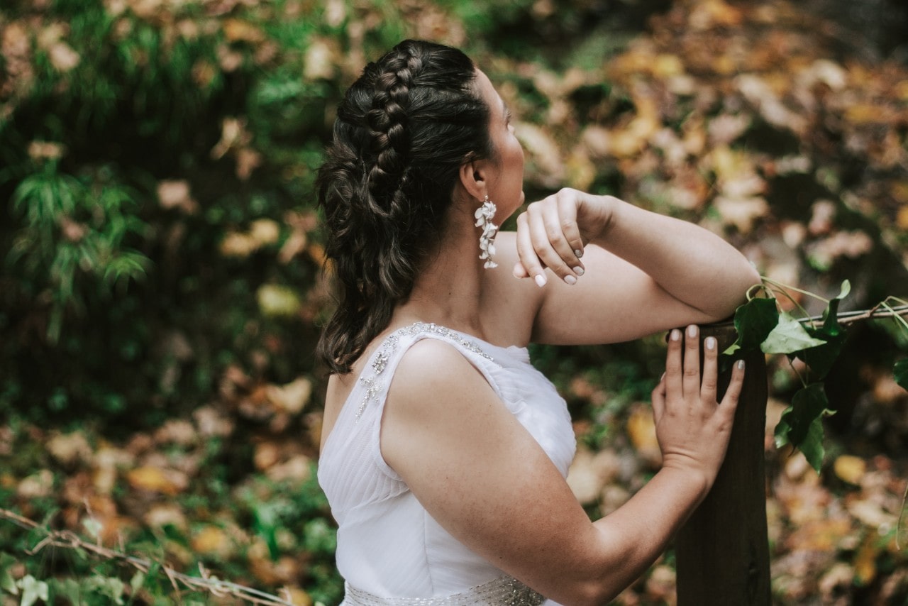 A bride looks off into the distance while leaning on a fence post.