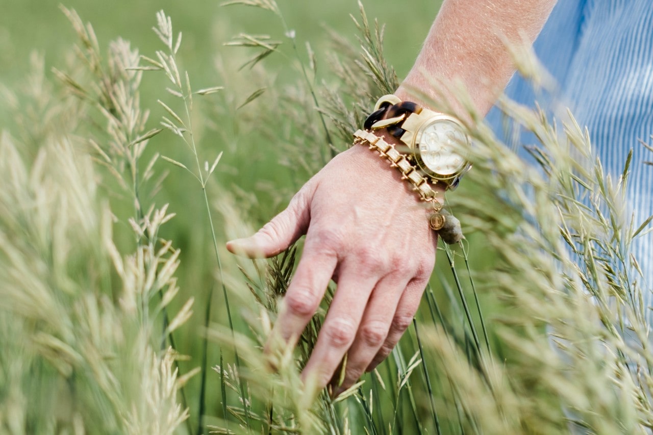 A close-up of a person’s hand wearing a bold watch and bracelet, brushing against tall grass in a field.