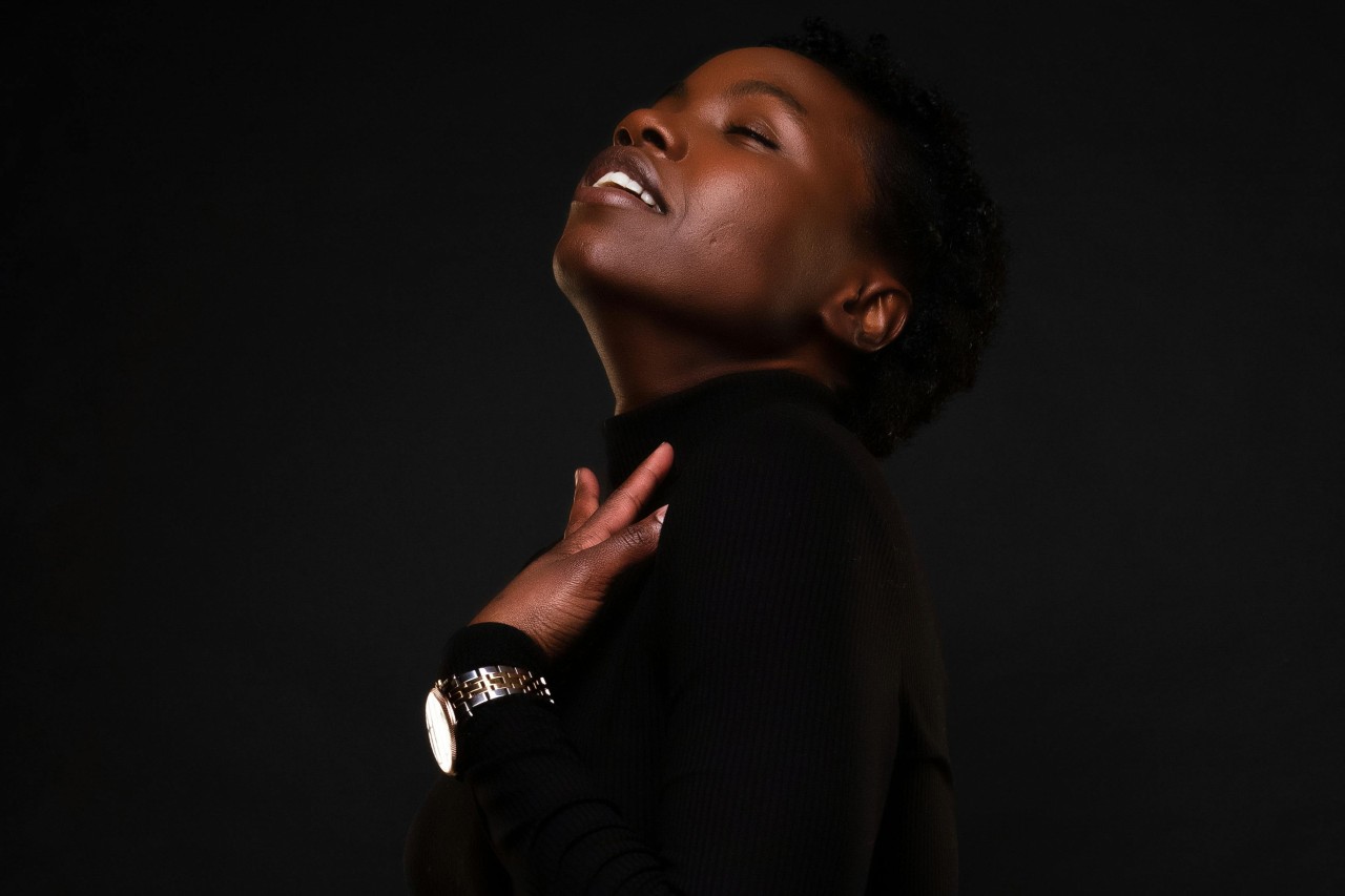 A close-up of a woman in a black sweater and large watch against a black background.