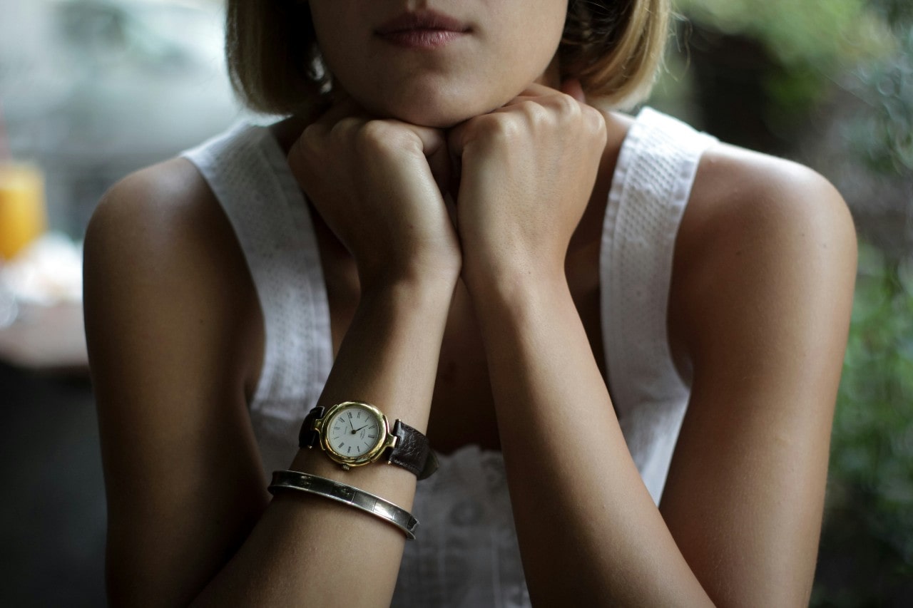 A woman wearing a gold watch with a round case, resting her chin on her hands.