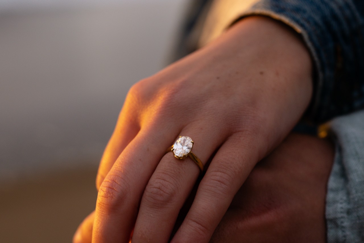 a woman’s hand resting on a man’s, wearing an oval cut engagement ring