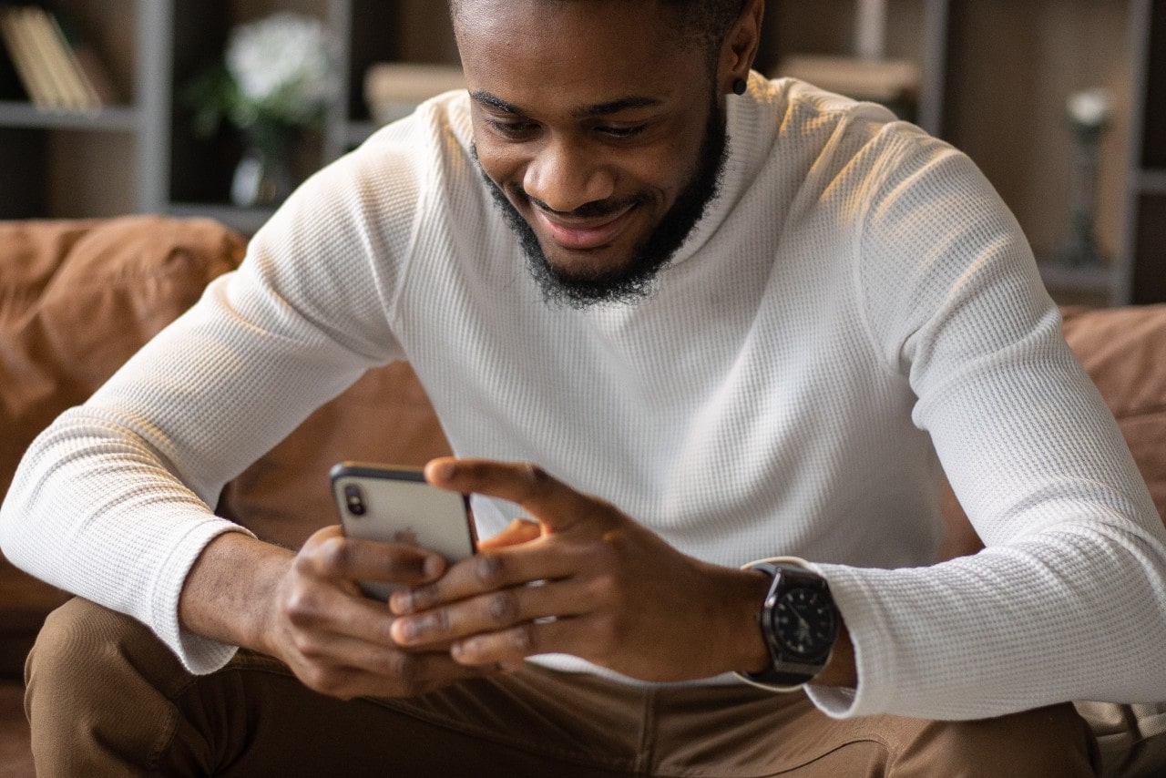 A man wearing a sports watch scrolls through his phone while sitting on a couch.