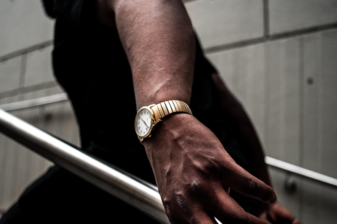 A man drags his wrist along a stair rail, wearing a gold watch.