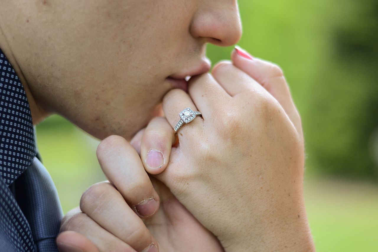 Cushion cut diamond set in an engagement ring with a diamond halo, accentuated by pave side stones. Worn on a woman’s hand pressed against her significant other’s chin.