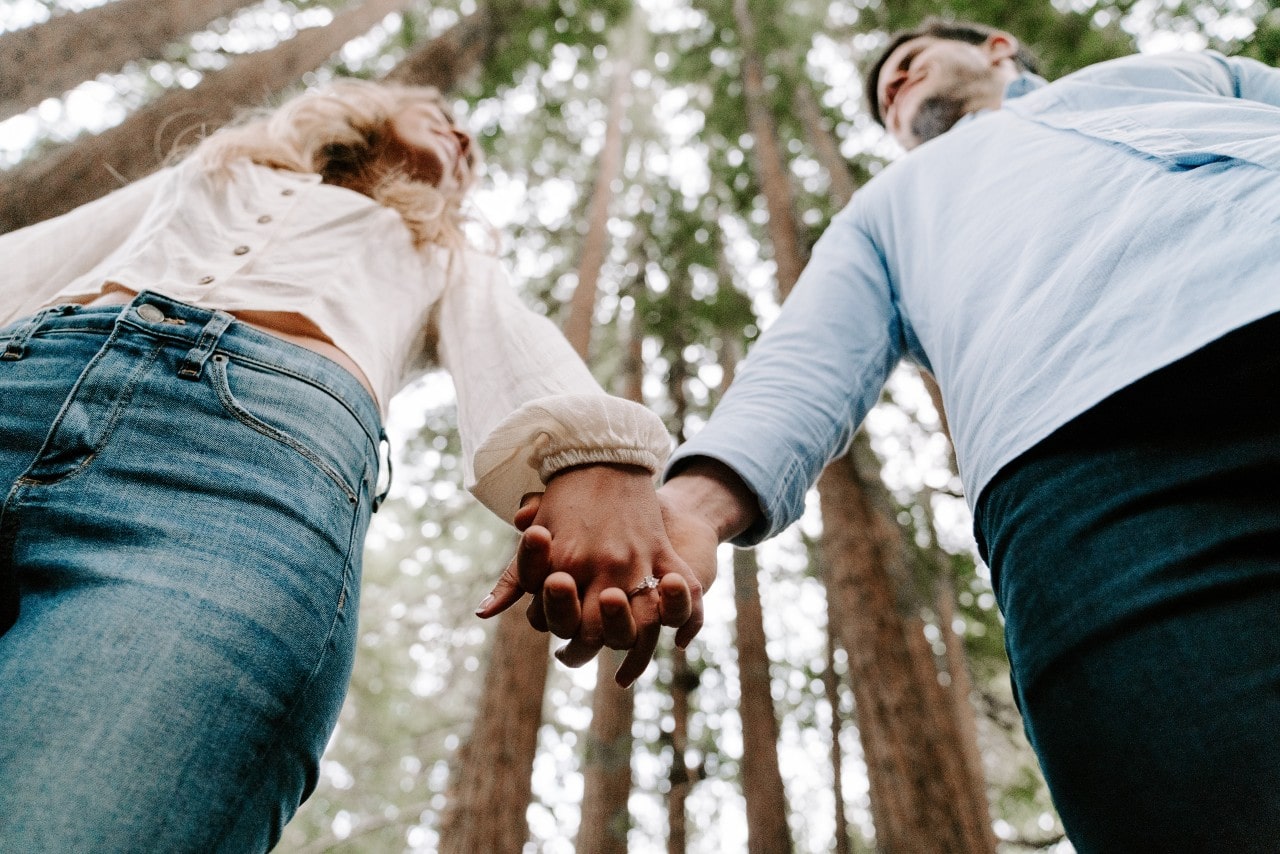 A low shot of a couple holding hands as they stroll through a wooded area, an engagement ring on the woman’s finger.