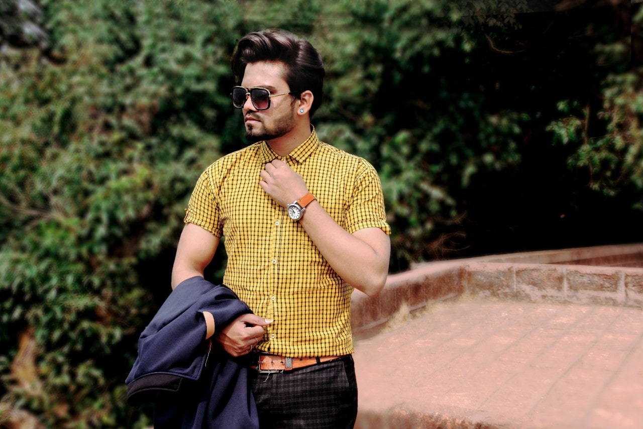 A man holds his sport jacket and adjusts his collar with a watch and sunglasses.