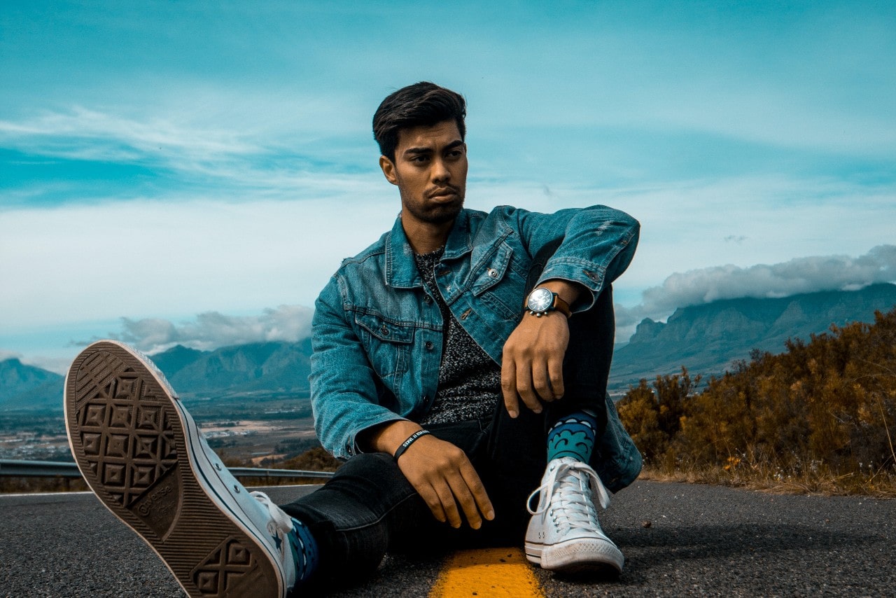 A man sits in the middle of a road, wearing Converse, a denim jacket, and a watch.